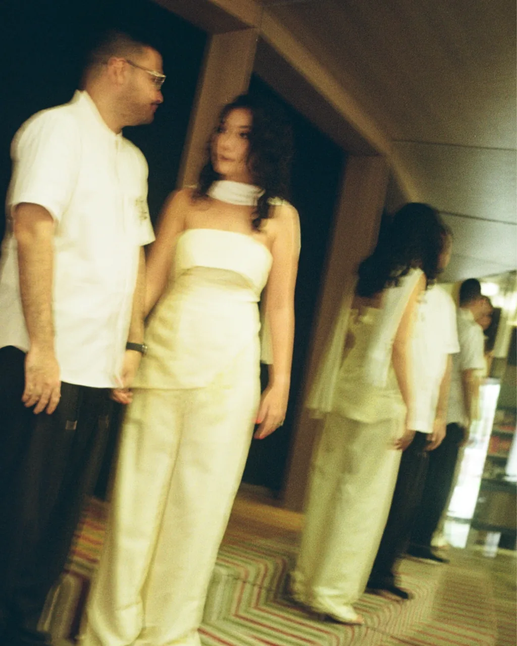 A couple in white and cream outfits standing close in a corridor, softly blurred by motion