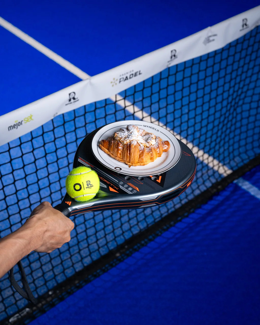 A croissant on a small plate balanced on a padel racket at the net, next to a tennis ball