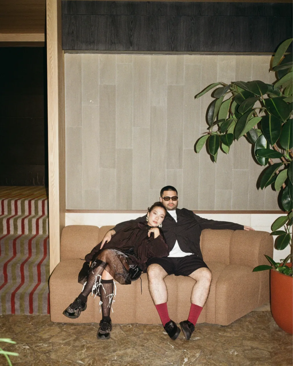 The couple in dark outfits resting together on a tan curved sofa beside a large plant, in front of a striped carpet