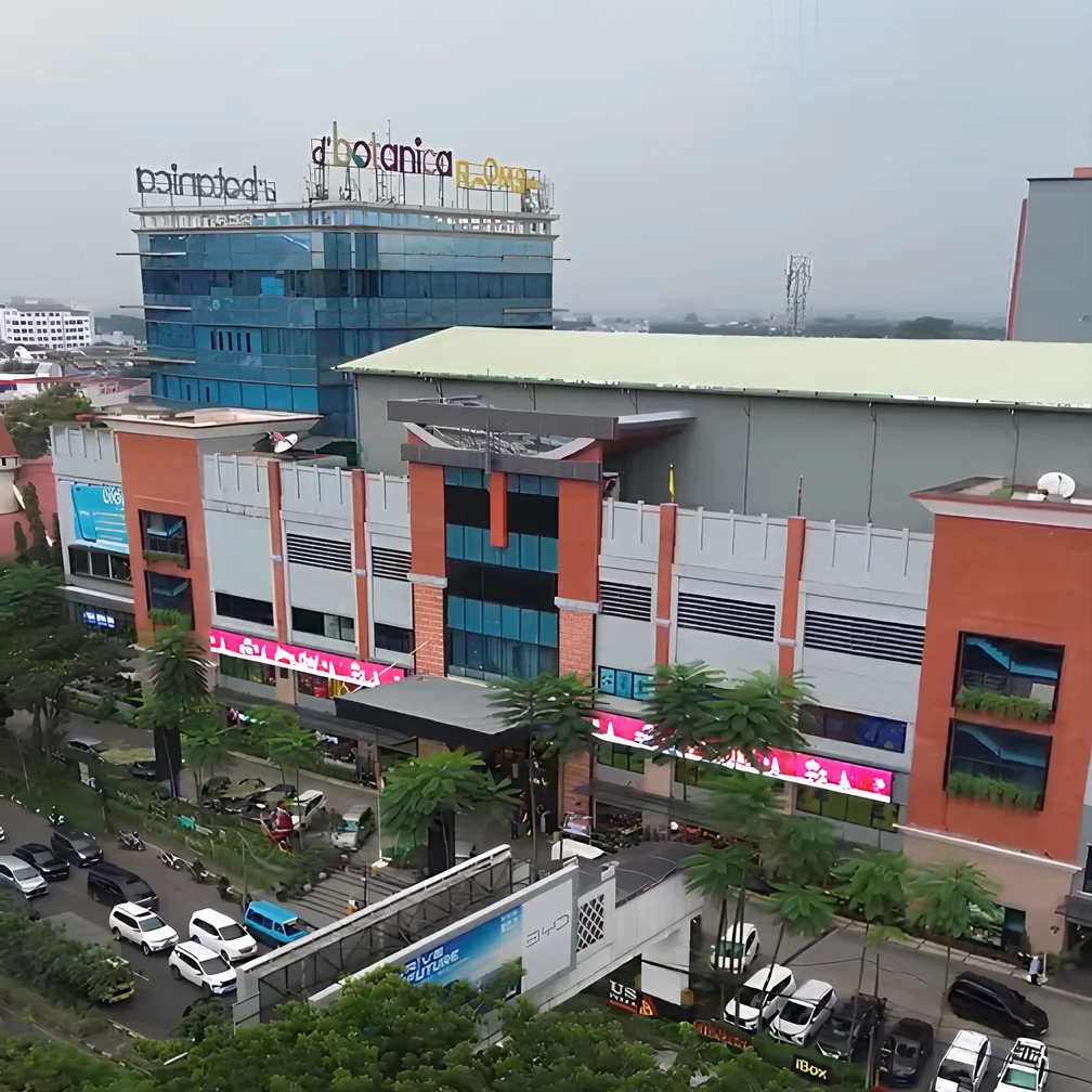 Aerial view of D'Botanica Bandung Mall: a blue glass tower with the d'botanica sign on top, and the brick-red mall facade with pink signage and palms in front