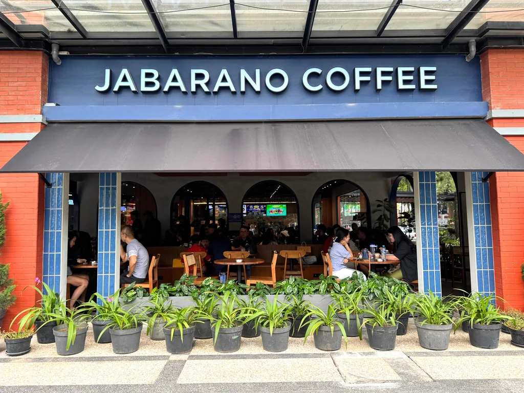 The Jabarano Coffee storefront at D'Botanica: a blue sign over a dark awning, red brick walls with blue tile columns, and guests at wooden tables behind a row of potted plants