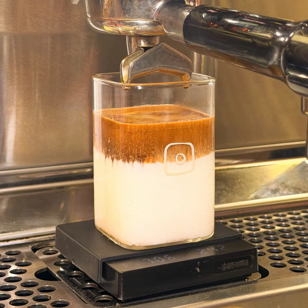 A square glass of milk with espresso poured on top, under the espresso machine, standing on a scale