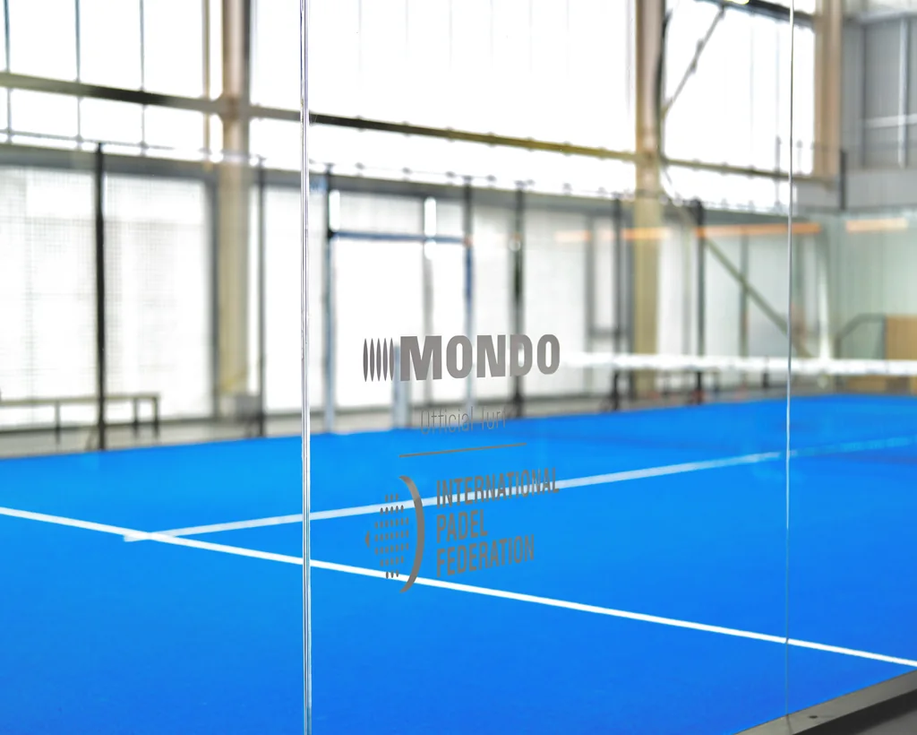 A blue padel court seen through a glass wall marked Mondo