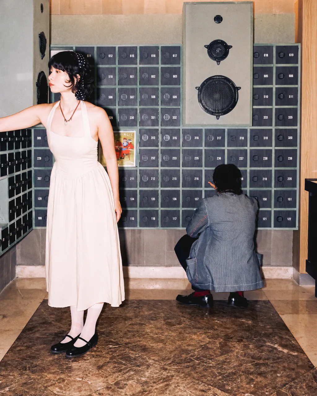 At the check-in counter: a woman in a white halter dress reaches toward the numbered key lockers while a man crouches at the lockers behind her