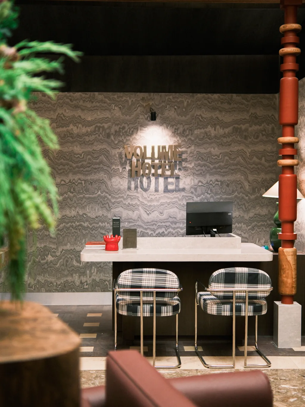 The front desk with the Volume Hotel logo on the wall, two checked bar stools and a red carved column